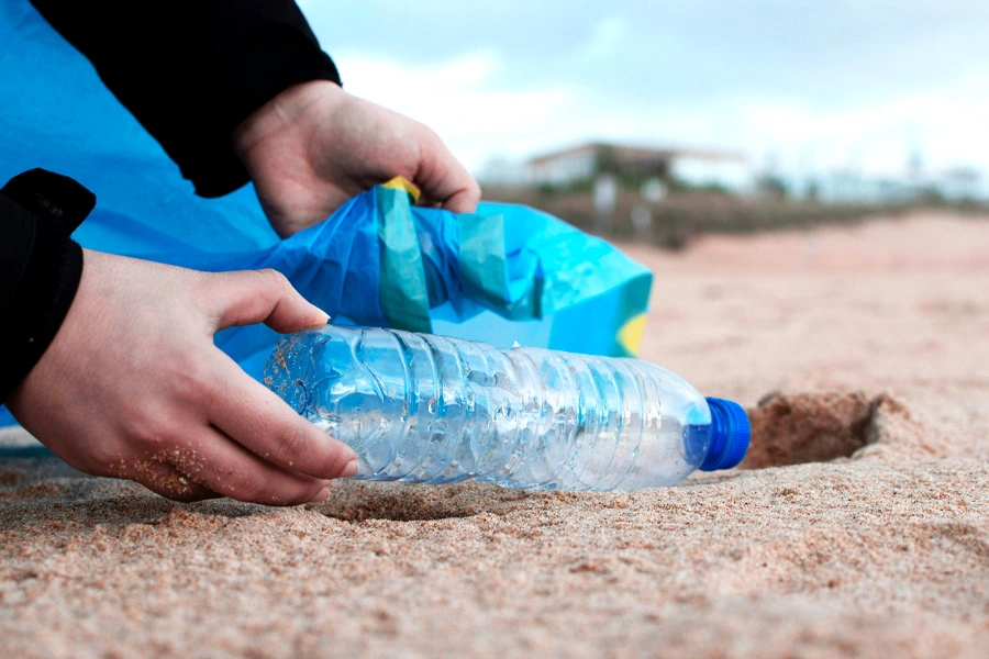 Uomo che raccoglie una bottiglia di plastica dalla sabbia e la mette in un sacco per la raccolta, a supporto della certificazione CSI di Vipitalia per plastica riciclata